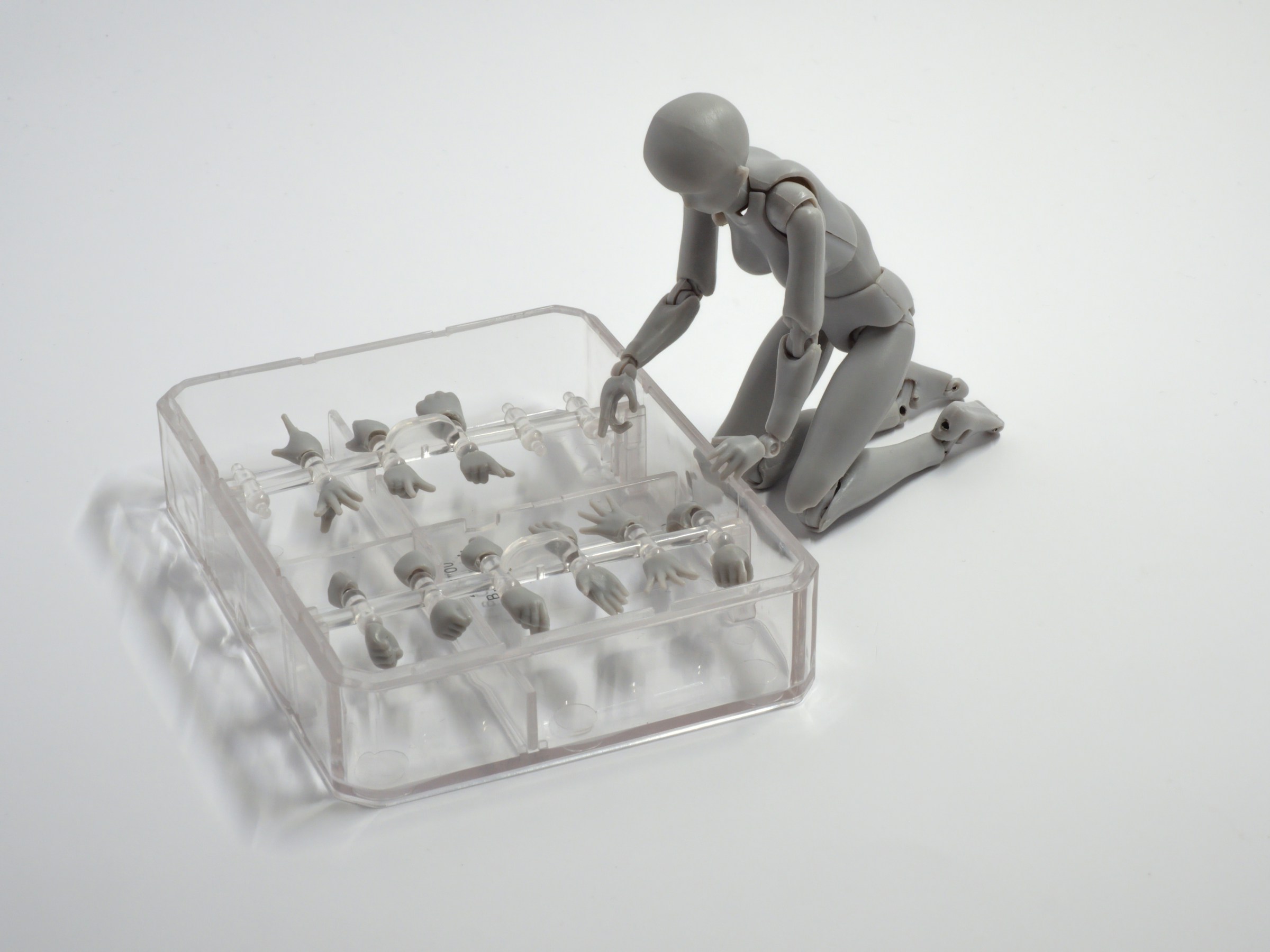 A grey poseable figurine kneels over an open case of interchangeable hands, choosing which pair to wear