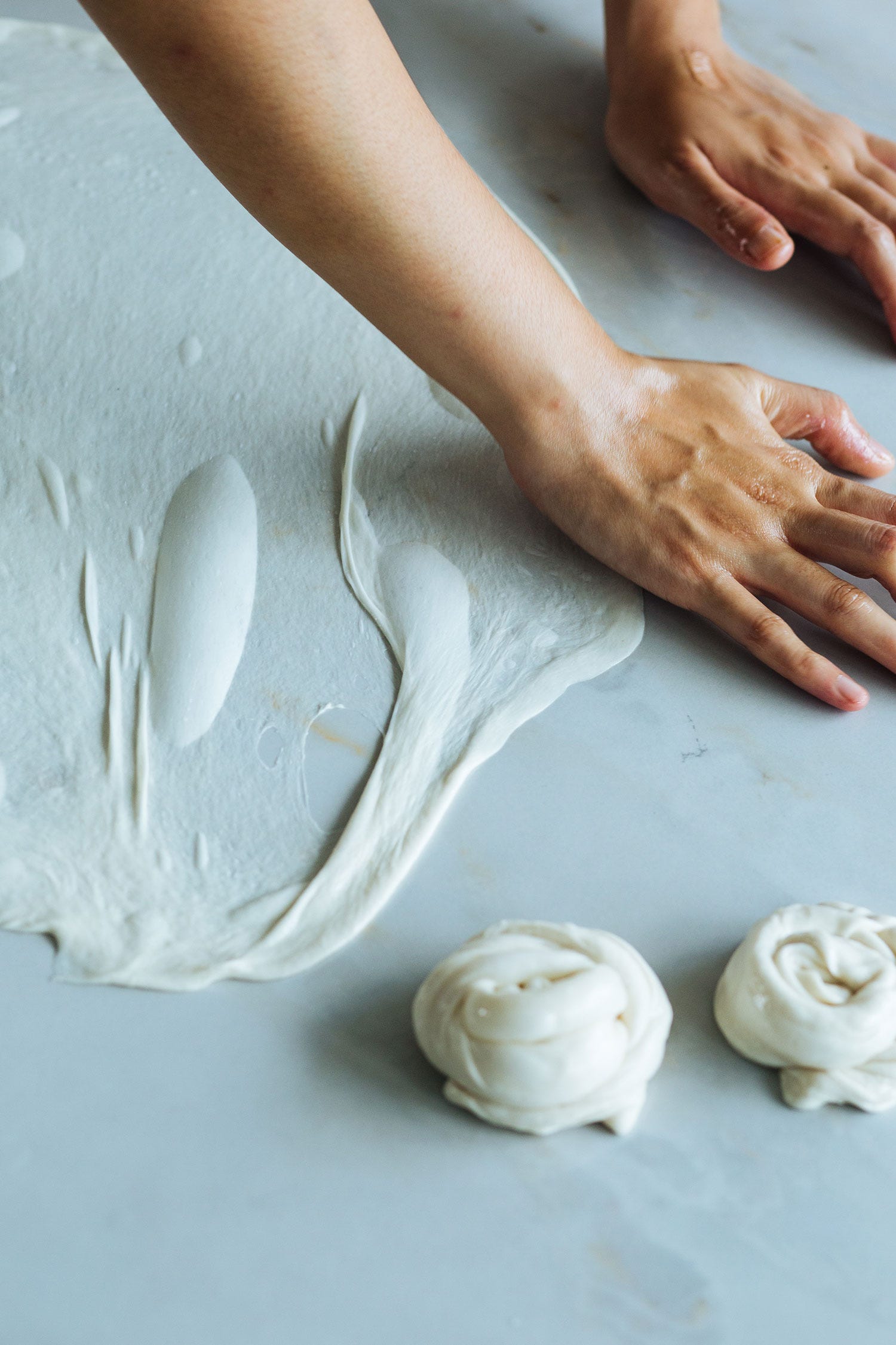 Hands stretching roti canai dough paper-thin on a marble counter, two coiled dough balls waiting beside it
